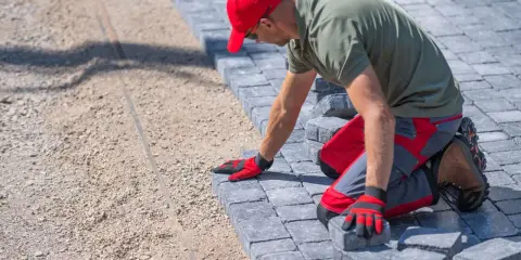 Pracownik w czerwonej czapce układa kostkę brukową. Poznaj ceny układania kostki brukowej i stwórz wymarzoną ścieżkę.