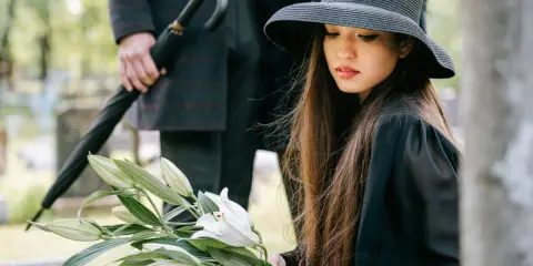 Kobieta w czarnym kapeluszu trzyma białe lilie na cmentarzu. Obok stoi mężczyzna z parasolem.
