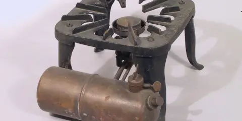 Gammalt fotogenkök i metall med en brännare och en bränsletank. Märkt "SVEA KOMFUR".