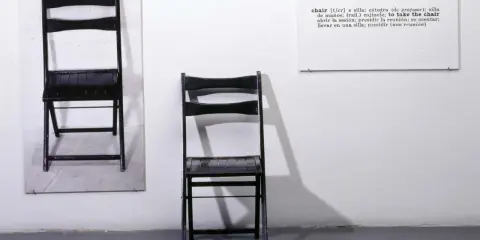 Una y tres sillas. Dos sillas negras plegables, una colgada en la pared y otra de pie, junto a un cartel con la definición de "chair".