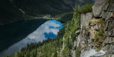 Gdzie jest Morskie Oko? Jezioro odbija chmury, otoczone lasem i górami. Wodospad spada po skalnej ścianie.