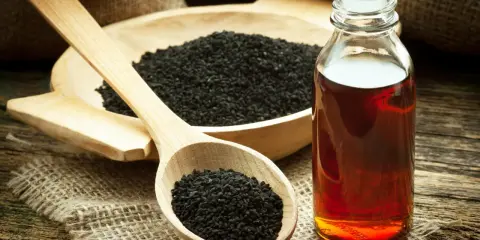 Huile de nigelle ambrée dans un flacon, à côté de graines noires dans une cuillère en bois. Découvrez les bienfaits de l'huile de nigelle.