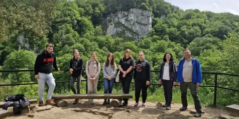Grupa studentów studiów ochrona środowiska podziwia malowniczy krajobraz z klifem i bujną zielenią.