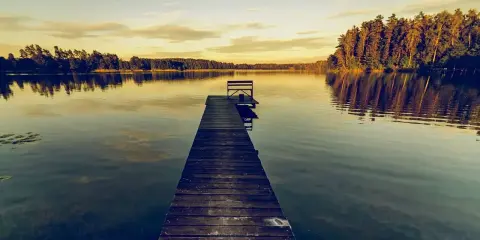 Drewniane molo z ławką na spokojnym jeziorze o zachodzie słońca, otoczone lasem.