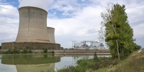 Kühltürme eines Atomkraftwerks in Frankreich, die Dampf abgeben. Ein ruhiger See spiegelt die Anlage wider, während am Ufer Bäume und Gras wachsen.