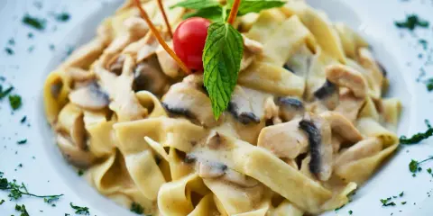 Pyszny makaron tagliatelle z kremowym sosem pieczarkowym, udekorowany pomidorkiem koktajlowym i listkiem mięty.