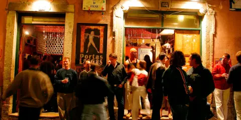 Menschen versammeln sich vor einem beleuchteten Lokal in Bairro Alto. Ein Mann hält ein Glas, andere unterhalten sich.