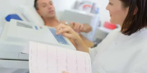 Pielęgniarka analizuje zapis EKG, który pokazuje, jakie są badania serca. Pacjent leży podłączony do aparatury.