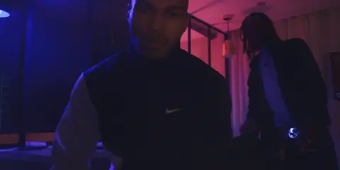 Dwóch mężczyzn w ciemnym pomieszczeniu, oświetlonym niebieskim i różowym światłem. Jeden z nich, 730 huncho, ma na sobie bluzę z logo Nike.