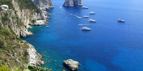 Scogliere verdi e rocce imponenti si affacciano sul mare blu intenso, con yacht che navigano. Un'immagine che ispira su come raggiungere Capri da Napoli.