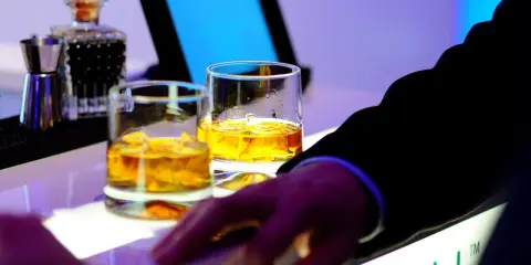 Dwa kieliszki złocistej whisky na barze, idealne na dzień whisky. Dłoń w garniturze sięga po drinka.
