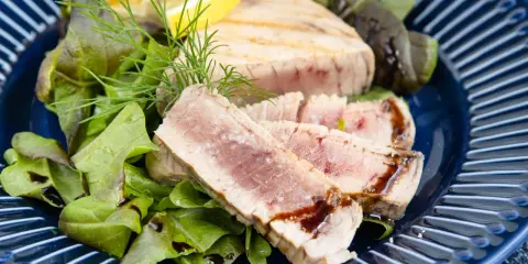 Ein perfekt gegrilltes Thunfischsteak, in Scheiben geschnitten, serviert mit frischem Salat, Zitrone und Dill auf einem blauen Teller.