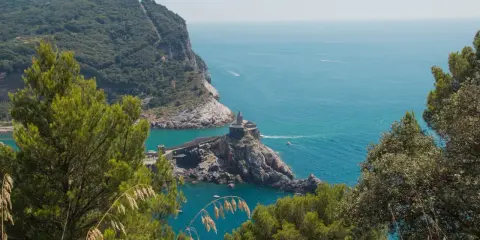 Vista panoramica del sentiero Riomaggiore Portovenere, con la chiesa di San Pietro su uno sperone roccioso e il mare azzurro.