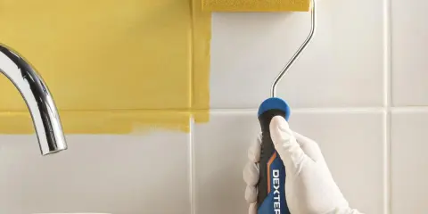 Mano con guante blanco pintando azulejos de cocina de blanco a amarillo con rodillo.