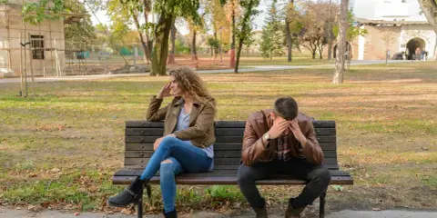 Pareja sentada en un banco, ella mira a un lado, él se tapa la cara. Parecen estar intentando procesar amor y desamor en un parque otoñal.