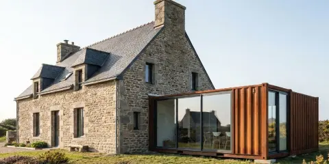 Maison en pierre avec une extension moderne en container maritime vitrée, offrant une vue sur la campagne fleurie.