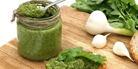 Słoik domowego pesto z rukoli, łyżka w środku, kromka chleba z pastą i świeże składniki: czosnek i rukola.