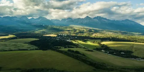 Malowniczy krajobraz z zielonymi polami i lasami, w oddali majestatyczne słowacja góry pod błękitnym niebem z chmurami.