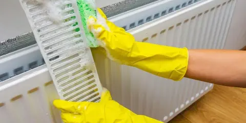Manos con guantes amarillos limpian la rejilla de un radiador con una esponja y espuma.