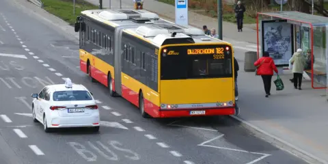Autobus linii 182 zbliża się do przystanku, a biała taksówka czeka, by ustąpić mu pierwszeństwa.
