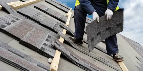 Dachówka bitumiczna układana na dachu. Pracownik w rękawiczkach trzyma kolejny element, przygotowując go do montażu.