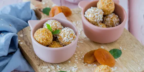 Zwei rosa Schalen gefüllt mit leckeren Energy Balls, bestreut mit Kokosraspeln und Haferflocken. Getrocknete Aprikosen und Minzblätter liegen daneben.