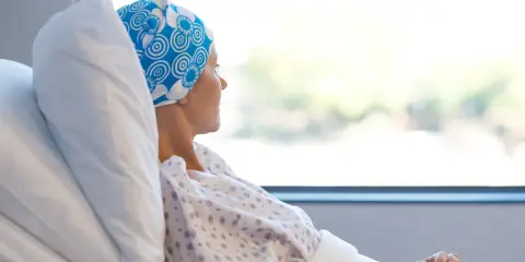Kobieta w chustce na głowie leży w szpitalnym łóżku, patrząc przez okno.