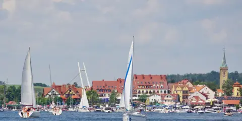 Piękne mazury widoki: żaglówki na jeziorze, miasteczko z czerwonymi dachami i wieżą kościelną w tle.