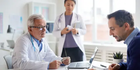 Lekarz omawia z pacjentem wyniki badań profilaktycznych dla mężczyzn. W tle asystuje pielęgniarka.