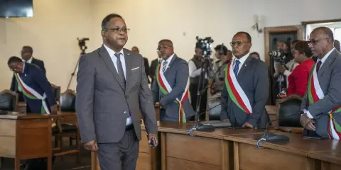 Des hommes en costume, certains portant des écharpes aux couleurs nationales, se tiennent dans une salle. L'un d'eux semble être une persona non grata, marchant à l'écart.