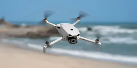 Biały dron DJI Lito X1 unosi się nad piaszczystą plażą, z morzem i niebem w tle.