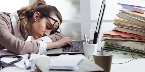 Zmęczona kobieta śpi na laptopie, otoczona stertami dokumentów. To obraz pracoholizmu, który doprowadził ją do wyczerpania.