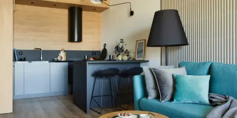 Nowoczesny salon z aneksem kuchennym 22m2. Turkusowa kanapa, czarne hokery przy wyspie, drewniane akcenty i stylowe oświetlenie tworzą przytulną przestrzeń.