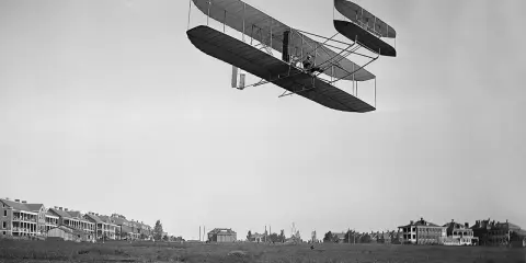 Das Kitty Hawk Flugzeug fliegt über ein Feld mit Gebäuden.