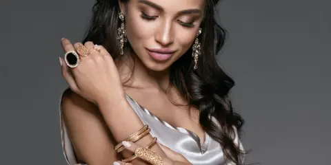 Kobieta w eleganckiej, satynowej sukience prezentuje bogatą biżuterię – złote bransolety, pierścionki z czarnymi kamieniami i kryształowe kolczyki. To kwintesencja glamour stylu ubioru.