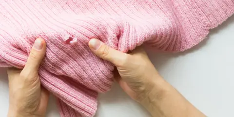 Dłonie trzymają różowy sweter z dziurą, pokazując problem, jak zaszyć dziurę w dresach.