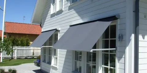 Grå markiser monterade på ett vitt trähus. Solen skiner och skapar skuggor på fasaden.