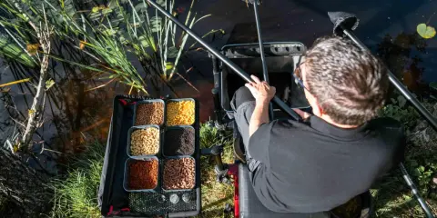 Wędkarz siedzi nad wodą, obok niego tacka z różnymi rodzajami przynęt, w tym gumki do pelletu.