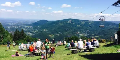 Widok na góry i miasto, ludzie odpoczywają na ławkach, w tle kolejka linowa. Bielsko-Biała atrakcje w pełnym słońcu.