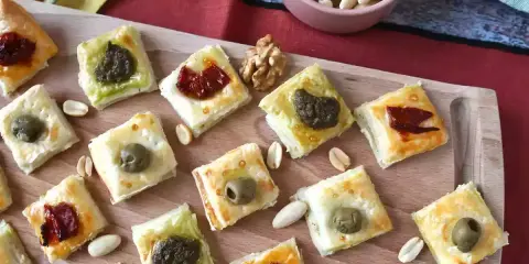 Rustici con pasta sfoglia, piccoli quadrati dorati con olive, pomodori secchi e pesto, disposti su un tagliere di legno.