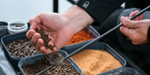 Ręka wsypuje granulki do pojemnika. W tle widać różne rodzaje zanęt, w tym otręby pszenne jak przygotować.