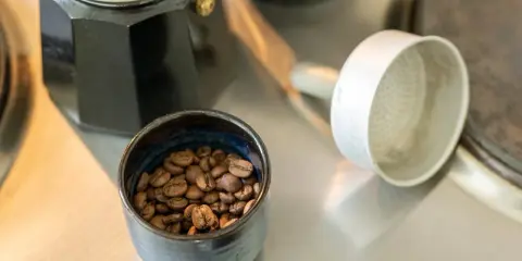 Kaffeebohnen in einer Schale neben einer Bialetti-Kaffeemaschine. Die Frage "bialetti wie viel kaffee" wird durch die Zubereitung beantwortet.
