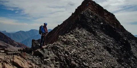 Alpinista na grani, zdobywający najwyższy szczyt Gruzji. W tle skaliste pasmo górskie i błękitne niebo.