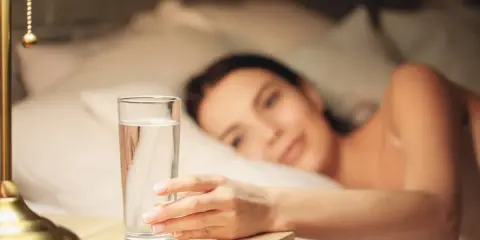 Une main attrape un verre d'eau posé à droite sur une table de chevet, une femme se réveille en arrière-plan.