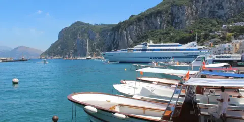 Barche in attesa al porto, con la maestosa isola di Capri sullo sfondo. Un traghetto Caremar attende passeggeri da Napoli.