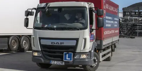 Biała ciężarówka DAF z naczepą, na której widnieje napis "Kategoria C1". Za kierownicą siedzi kierowca w masce.