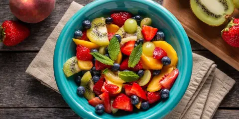 Un bol turquoise débordant de salade de fruits été : fraises, myrtilles, kiwis, pêches et raisins, garnie de menthe fraîche.