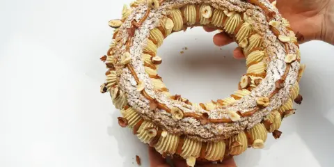 Un Paris-Brest craquelin, orné de noisettes et de caramel, est tenu par des mains.