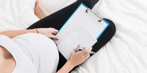 Kobieta w ciąży leży na łóżku z notatnikiem i długopisem, zastanawiając się, czy na macierzyńskim można pracować.