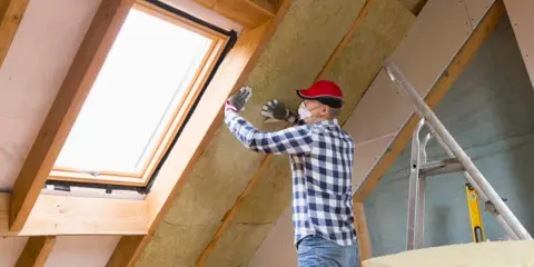 Mann isoliert ausgebauten Dachboden mit Dämmmaterial. Ein Dachfenster sorgt für Licht.
